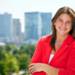 Headshot of Amanda Karfakis, CEO of the branding-digital-PR agency Vitamin standing on the rooftop of the agency's headquarters at 720 Light Street in Baltimore, Maryland, with the cityscape buildings and trees as a backdrop.