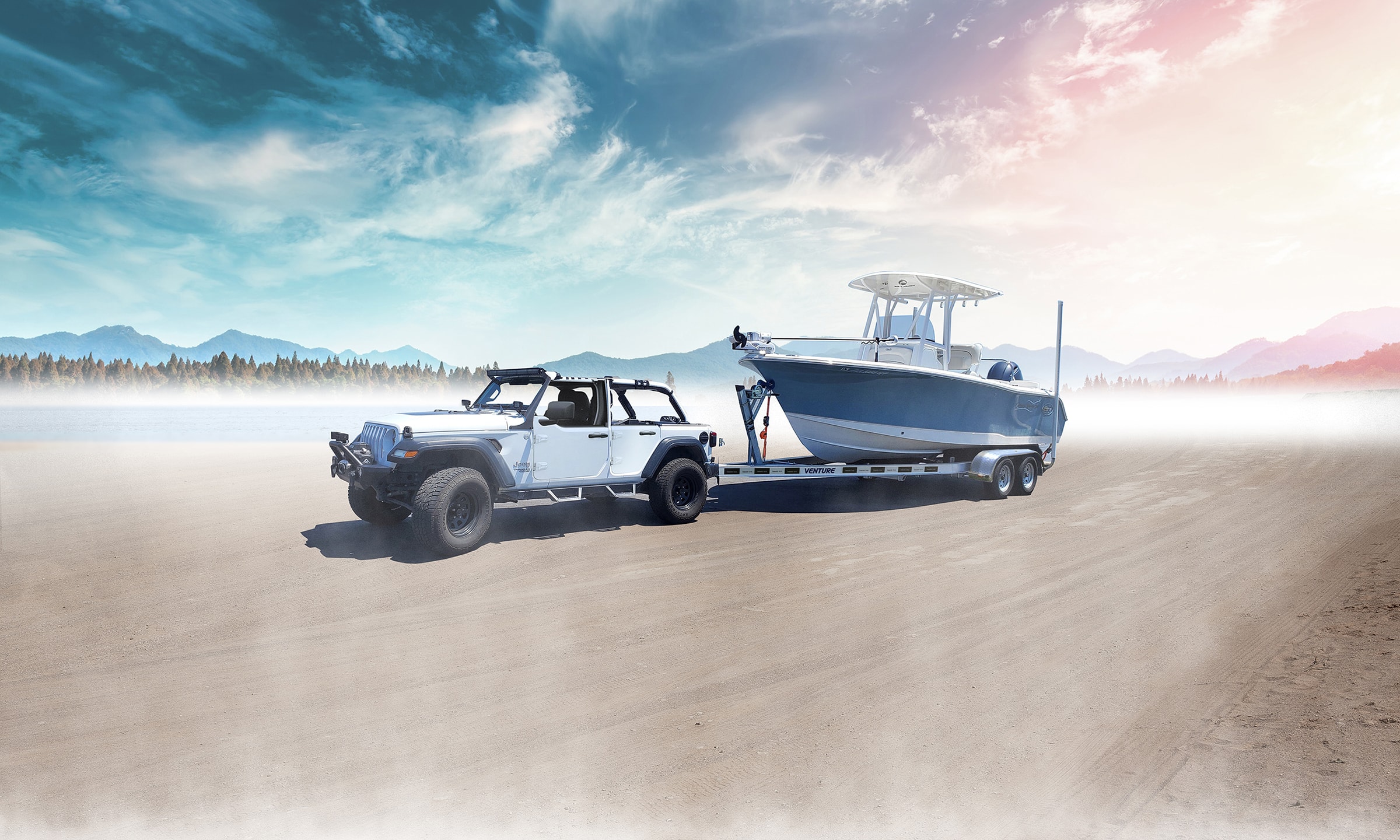 Photograph of a Jeep towing a boat on a Venture Trailers boat trailer along a sandy road, with a mountainous skyline and blue cloudy skies in the background